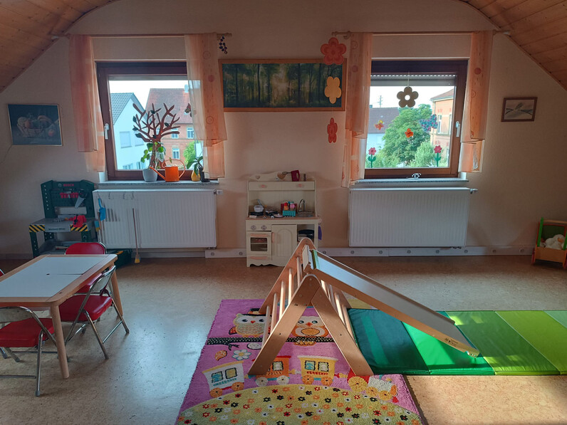 Bild: Spielzimmer mit einem kleinen Kindertisch mit drei Stühlen und einer kleinen Rutsche auf Matten. Im Hintergrund zwischen zwei Fenstern eine Spielküche aus Holz. Ganz links eine kleine grüne Spielwerkbank.